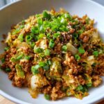 Close-up of a hearty serving of egg roll in a bowl with ground meat, cabbage, topped with fresh green onions and sesame seeds.