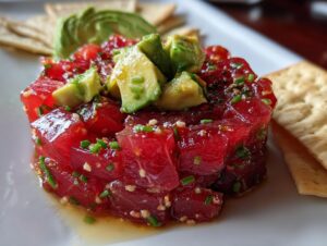 Close-up of a perfectly molded Tuna Tartare Recipe, topped with diced avocado and served with crackers.