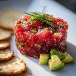 A perfectly molded stack of bright red Tuna Tartare Recipe garnished with chives and sesame seeds, served with avocado cubes and crackers.
