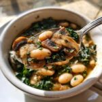A spoonful lifted from a bowl showing white bean mushroom stew with kale and fresh herbs.