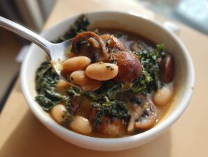 A spoonful of rich white bean mushroom stew featuring white beans, dark mushrooms, and green kale.