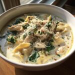 A close-up of a white bowl filled with creamy white chicken lasagna soup, topped with shredded chicken, spinach, and Parmesan cheese.