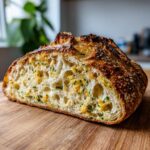 A close-up of a freshly baked artisan bread loaf, sliced open to show the open crumb filled with yellow/orange filling and green herbs.