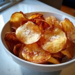 A white bowl filled with golden brown, crispy air fryer potato chips sprinkled with coarse sea salt.