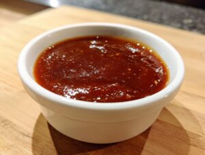 Close-up of rich, dark red homemade bbq sauce glistening in a small white ramekin on a wooden board.