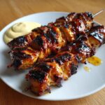 Close-up of perfectly grilled, glazed chicken skewers served with a side of yellow dipping sauce.