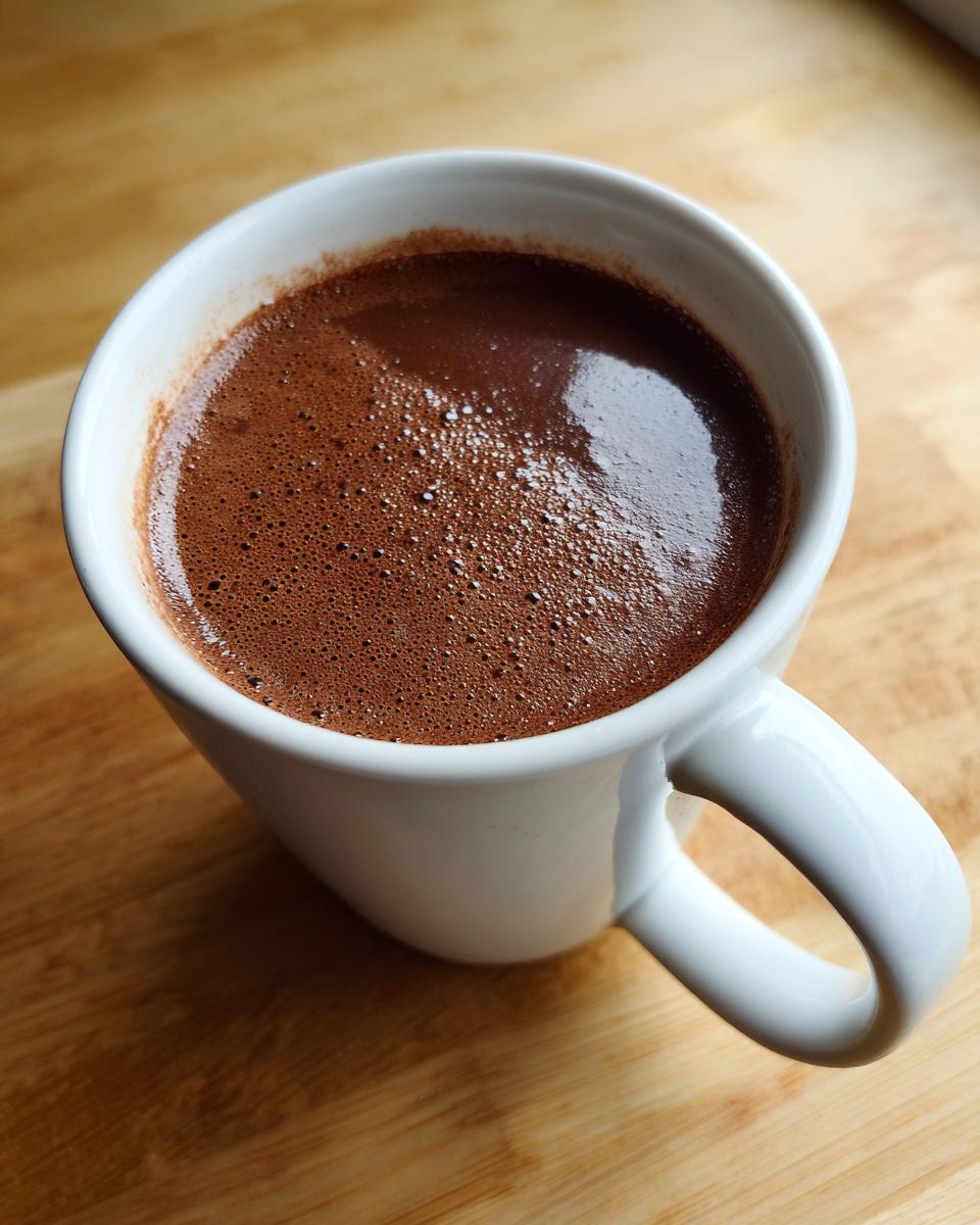 Overhead view of a steaming white mug filled with rich, dark hot cocoa mix resting on a wooden surface.