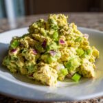 A mound of creamy avocado chicken salad mixed with chunks of avocado, red onion, and green onion served on a white plate.