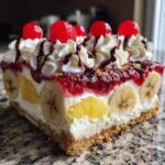 Close-up of a slice of banana split cake showing layers of crust, cream, bananas, pineapple, strawberry topping, whipped cream, and cherries.