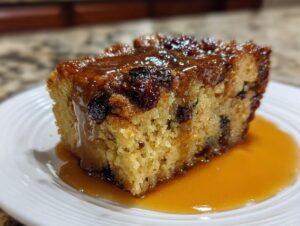 A close-up of a moist slice of bread pudding with bourbon sauce drizzled over raisins and pooling on a white plate.