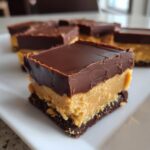 A square piece of buckeye fudge bar showing distinct layers: chocolate topping, thick peanut butter filling, and a dark crust.