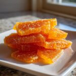 A close-up stack of bright orange, sugary candied orange slices resting on a white square plate.