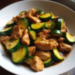 A close-up of savory chicken zucchini stir fry pieces coated in a glossy brown sauce served in a white bowl.