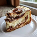 A generous slice of cinnamon roll cheesecake showing layers of cake, cinnamon swirl, and creamy cheesecake filling, topped with icing.