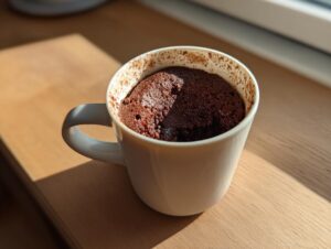 A freshly baked Cottage Cheese Microwave Brownie puffed up inside a white ceramic mug, sitting on a wooden surface.