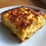 A square slice of golden brown cream cheese corn casserole with visible kernels on a white plate.