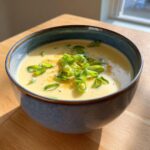 A close-up of creamy cauliflower soup in a blue bowl, garnished with fresh green scallions and black pepper.