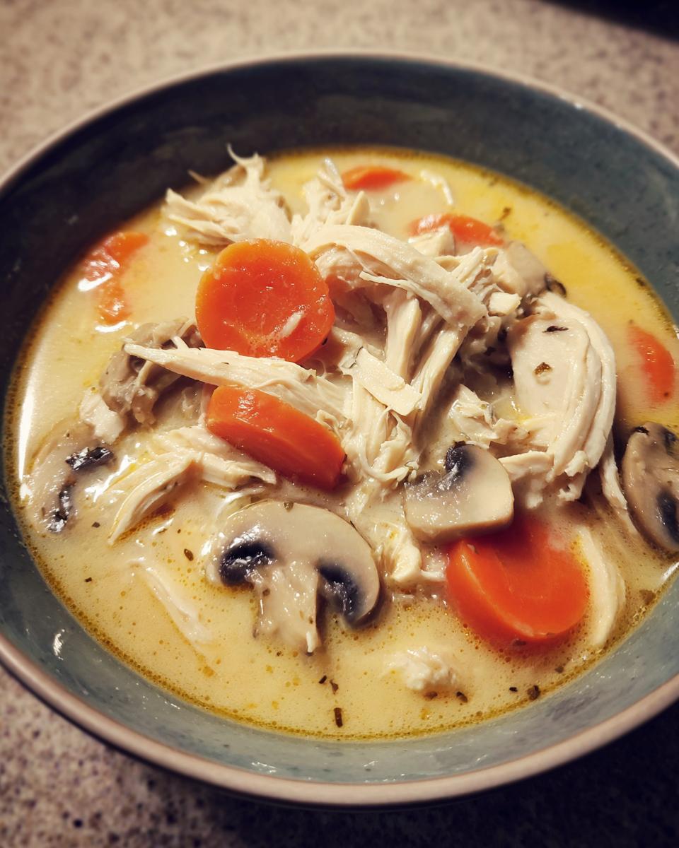 A close-up of a bowl of creamy coconut soup filled with shredded chicken, sliced carrots, and mushrooms.