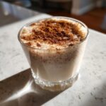 A close-up of a glass filled with creamy coquito, topped generously with ground cinnamon.