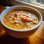 Close-up of a thick, creamy bowl of navy bean soup topped with savory ham pieces and black pepper.