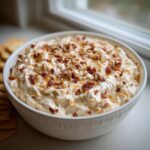 A white bowl filled with creamy Neiman Marcus dip, topped generously with chopped pecans and bacon bits.