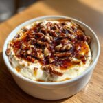 A close-up of a creamy pecan pie dip in a white bowl, generously topped with candied pecans and caramel sauce.