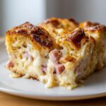 Close-up of a cheesy Croissant Breakfast Casserole slice showing flaky layers, ham chunks, and melted white sauce.
