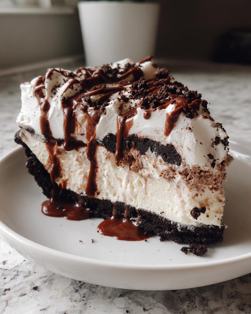 A tall slice of layered ice cream pie with a dark crust, creamy filling, whipped topping, and chocolate drizzle.