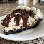 A close-up of a rich slice of ice cream pie with a dark crust, creamy filling, whipped topping, chocolate drizzle, and cookie crumbs.