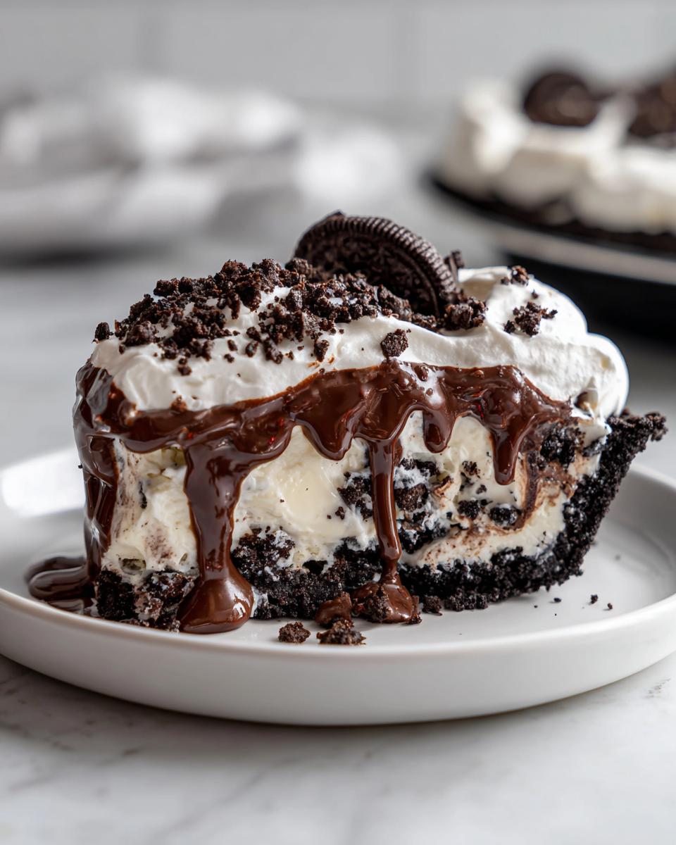 Close-up of a rich slice of ice cream pie with a dark cookie crust, vanilla filling, whipped cream, and dripping chocolate fudge.