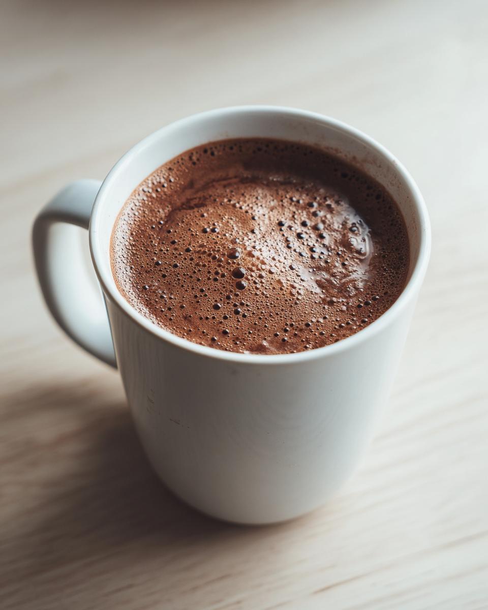 Close-up of a white mug filled with freshly made, foamy hot cocoa mix resting on a light wooden surface.
