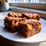 Four rich, fudgy apple brownies with visible chunks of apple served on a white plate near a window.