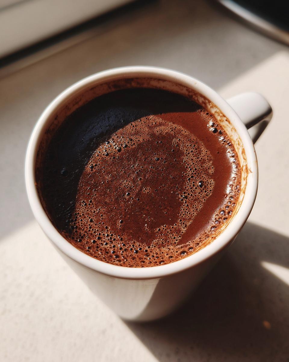 Close-up of rich, dark french hot chocolate with a foamy top in a white mug, illuminated by sunlight.