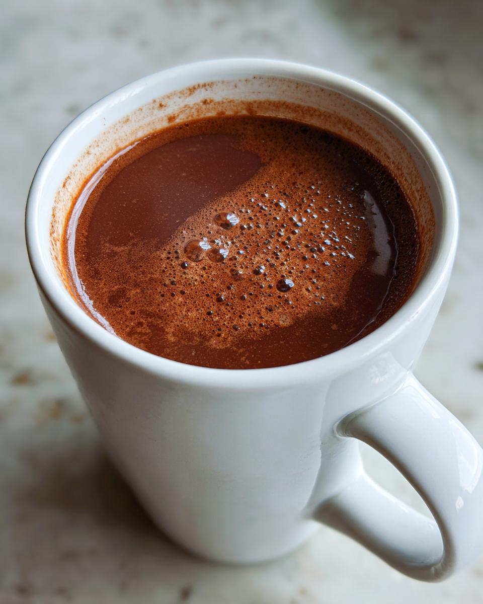 Close-up of rich, dark french hot chocolate with a slight foam layer in a white ceramic mug.