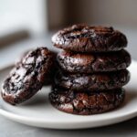 A stack of four rich, dark brownie cookies with shiny, crinkled tops, plus one leaning against the stack.