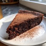 A rich, dark slice of fudgy flourless chocolate torte dusted generously with cocoa powder on a white plate.