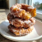 Three golden brown baked apple fritters stacked on a white plate, drizzled generously with white glaze.