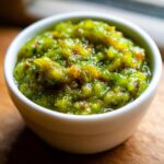 A close-up, bright shot of chunky green tomato salsa served in a small white bowl on a wooden surface.