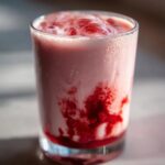 A tall glass filled with homemade strawberry milk showing swirls of bright red strawberry syrup mixing with the pink liquid.