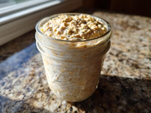 A glass jar filled with creamy, textured overnight oats sitting on a granite countertop near a window.