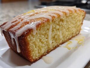 Close-up of a moist slice from a lemon cake recipe, topped with thick white lemon glaze drizzling down the sides.
