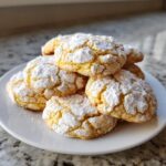 A stack of bright yellow lemon crinkle cookies generously dusted with powdered sugar, resting on a white plate.
