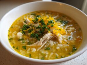 Close-up of Lemon Ginger Turmeric Chicken and Rice Soup, featuring shredded chicken, rice, and yellow broth, topped with fresh parsley and zest.