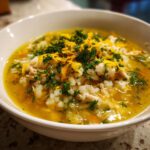 Close-up of a white bowl filled with Lemon Ginger Turmeric Chicken and Rice Soup, topped with shredded chicken, rice, parsley, and zest.