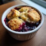 Close-up of a warm mixed berry cobbler with golden biscuit topping bubbling over dark purple berries in a white ramekin.