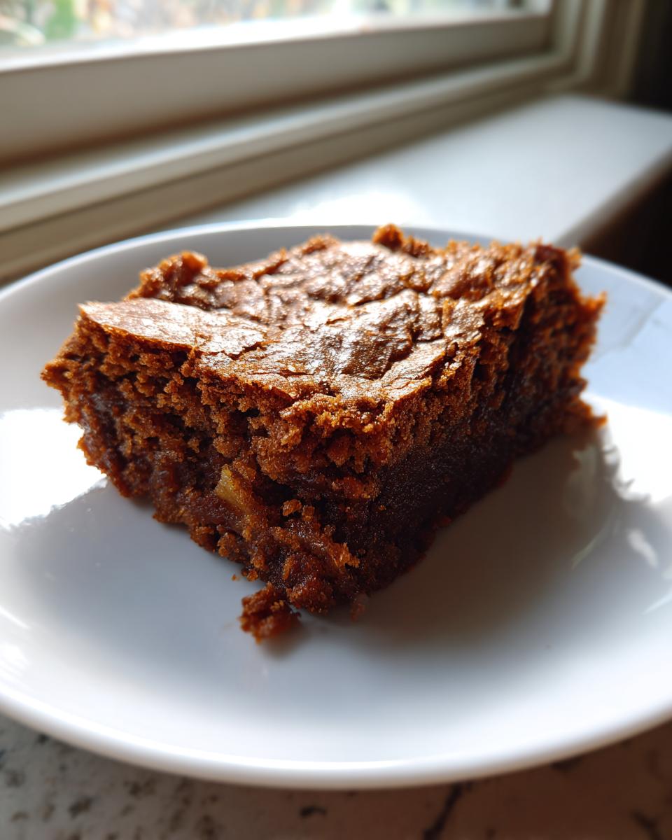 A close-up of a rich, dark, moist square of apple brownies resting on a white plate near a window.