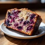 Close-up of a moist slice of blueberry banana bread packed with dark purple blueberries on a small white plate.
