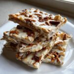 A stack of squares of homemade peanut butter pretzel bark topped with white chocolate and nuts on a white plate.