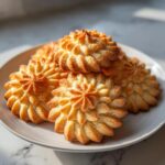 A stack of beautifully piped, golden brown butter cookies shaped like flowers on a light plate.