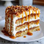 A tall slice of pumpkin poke cake layered with white frosting, drizzled heavily with caramel sauce and topped with nuts.
